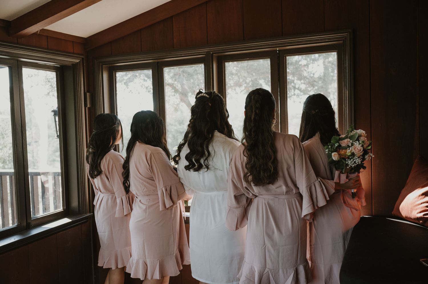 Bride and bridesmaids in white robes standing together looking out wooden cabin windows before wedding ceremony.
