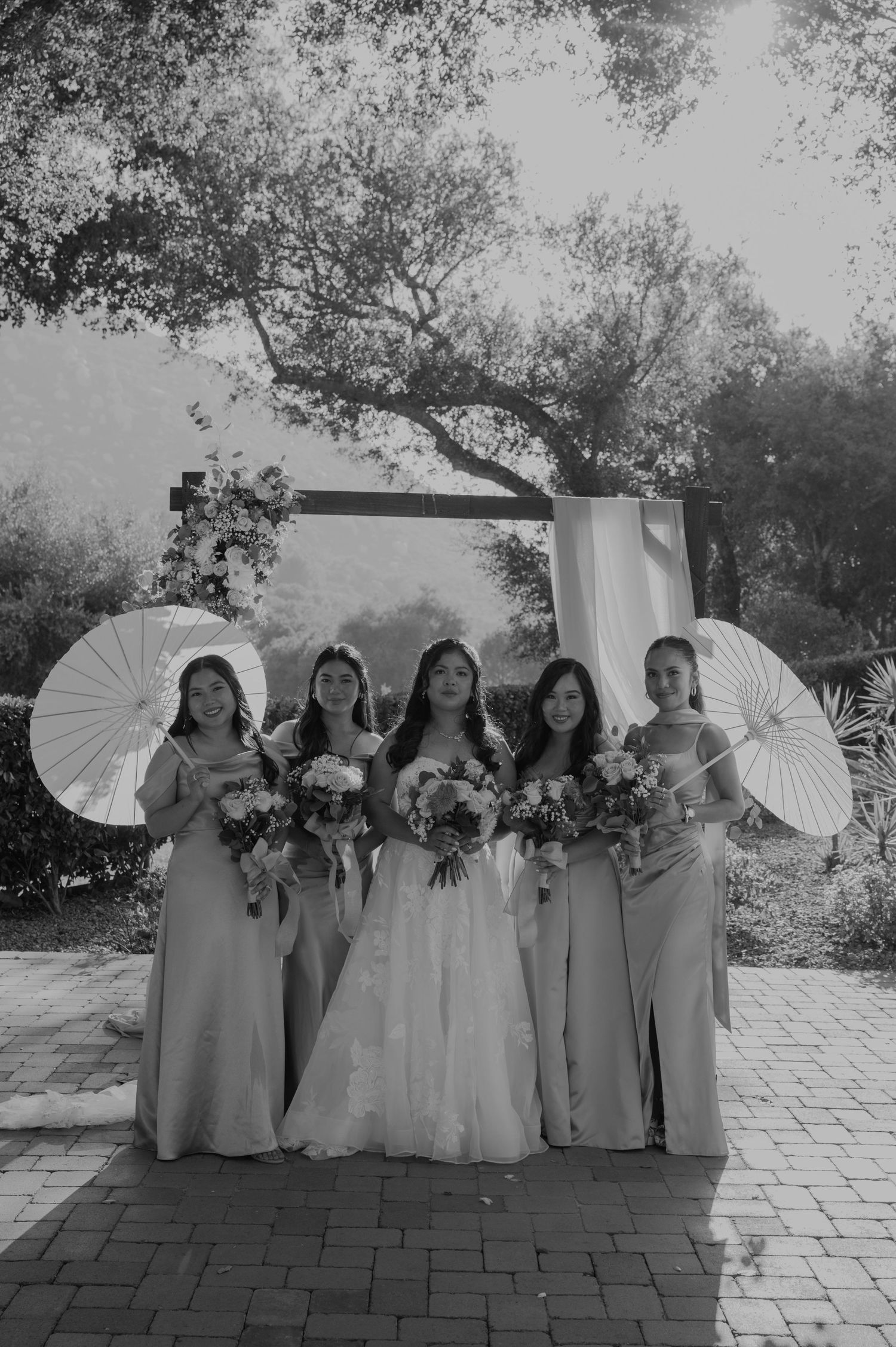 Bride and four bridesmaids holding bouquets and parasols in black and white photo before wedding arch.