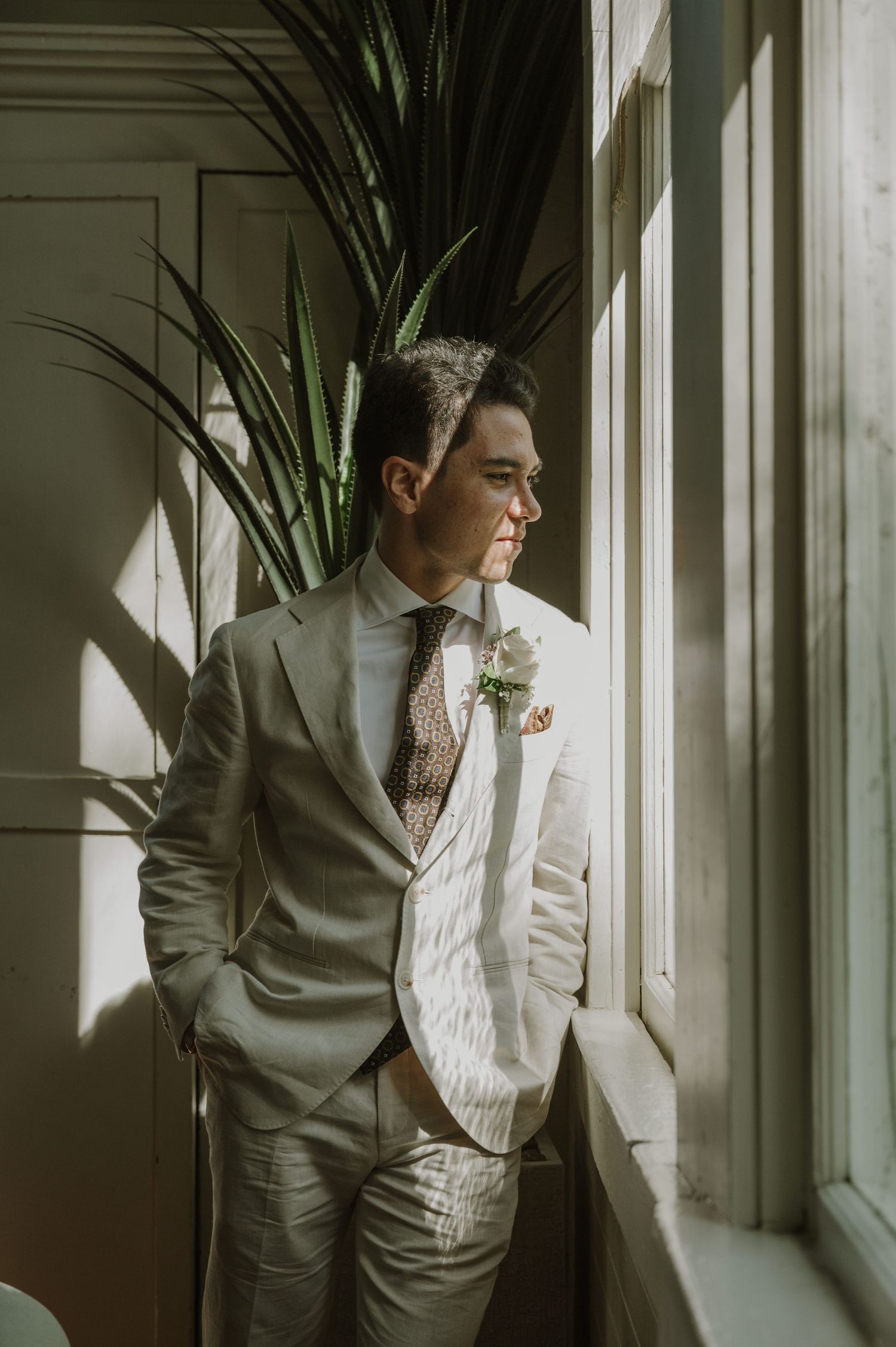 Elegant groom in light linen suit with boutonniere gazing out sunlit window beside tall green plant.