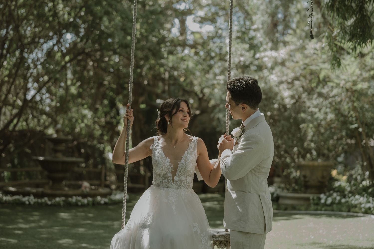 Bride in lace gown sits on rope swing as groom in gray suit holds her hands in a lush garden wedding setting.