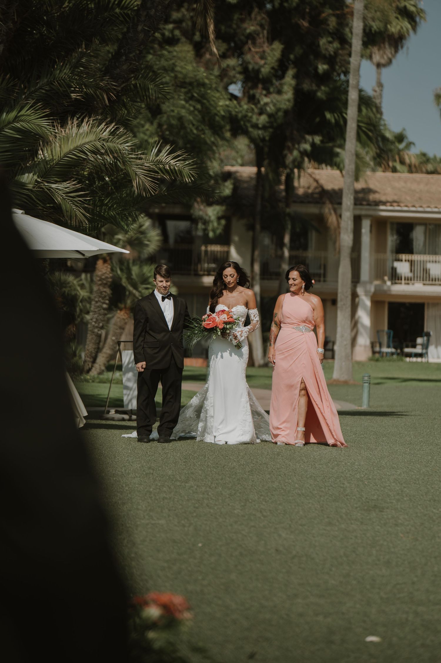 Wedding party members walk across a manicured lawn with palm trees at an elegant outdoor venue.