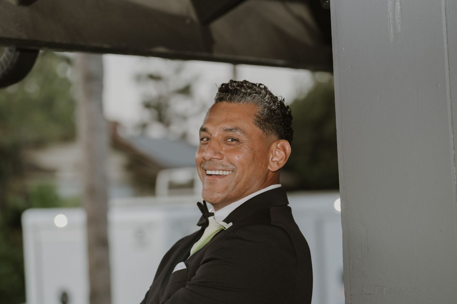 Smiling man in black tuxedo with green tie, photographed outdoors at a formal event.