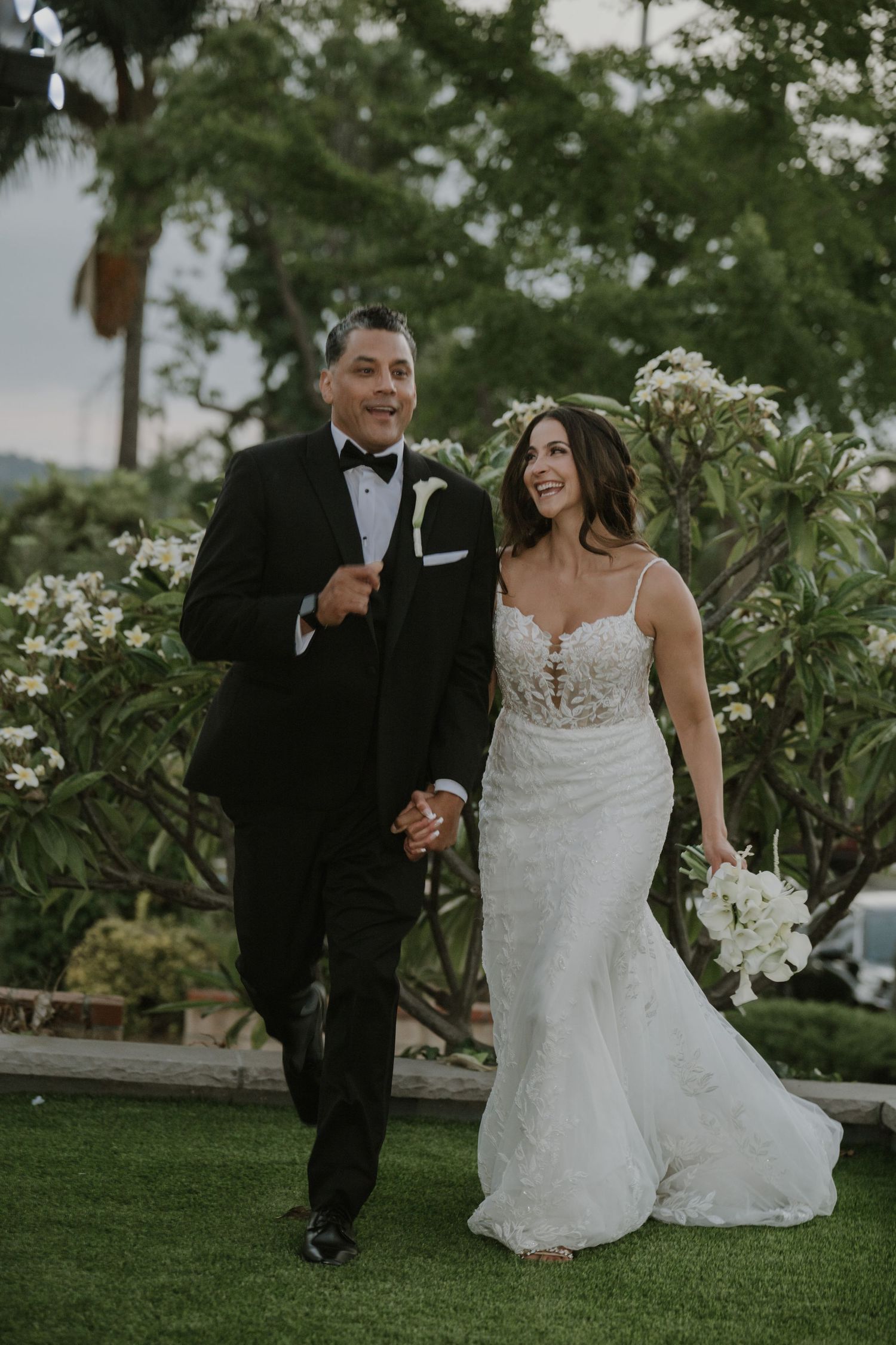 Happy bride and groom walking hand-in-hand on green lawn, bride in lace gown holding white bouquet.