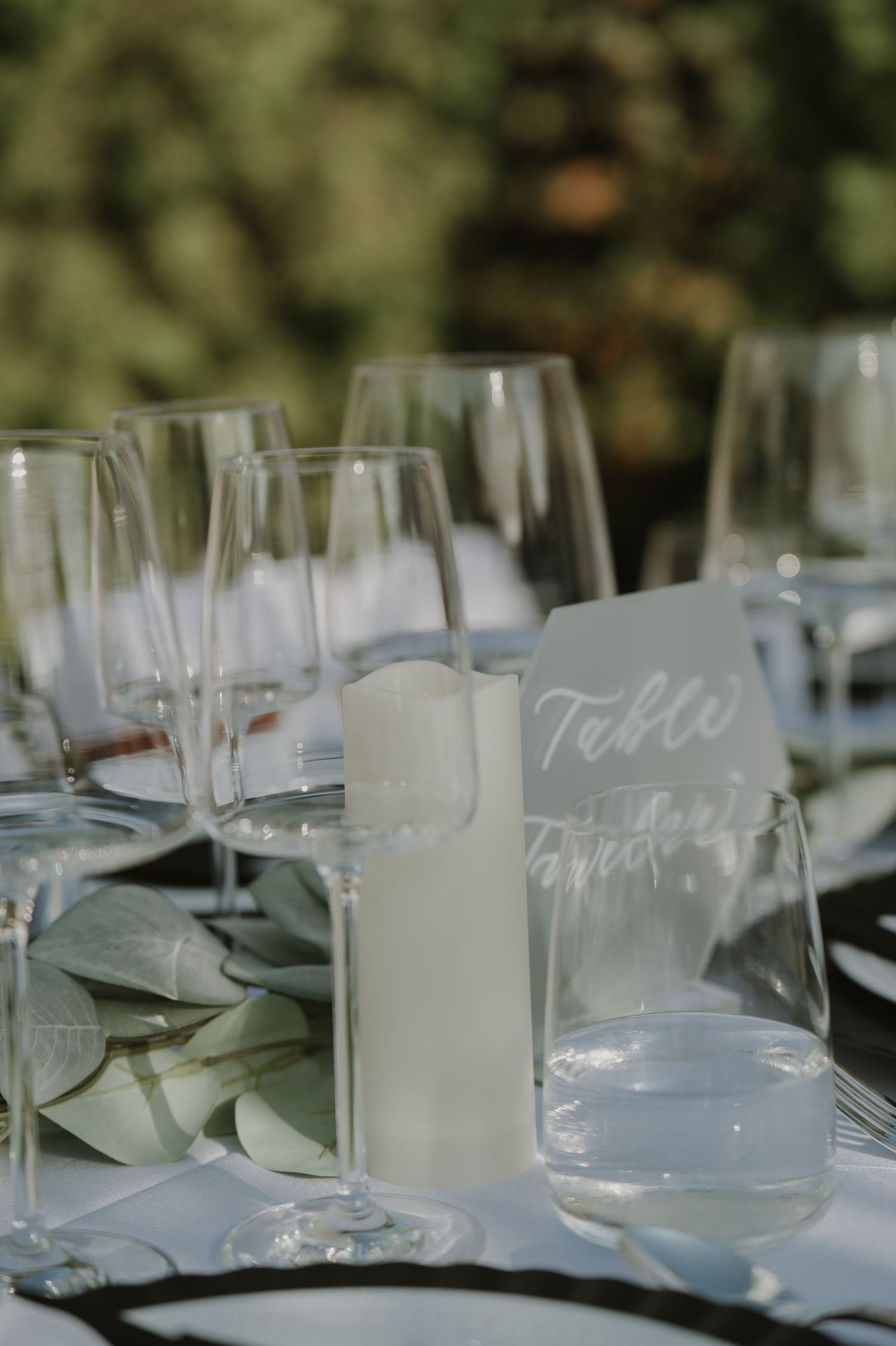 Outdoor wedding table setting with stemless wine glasses, white candle, and calligraphy table number card.