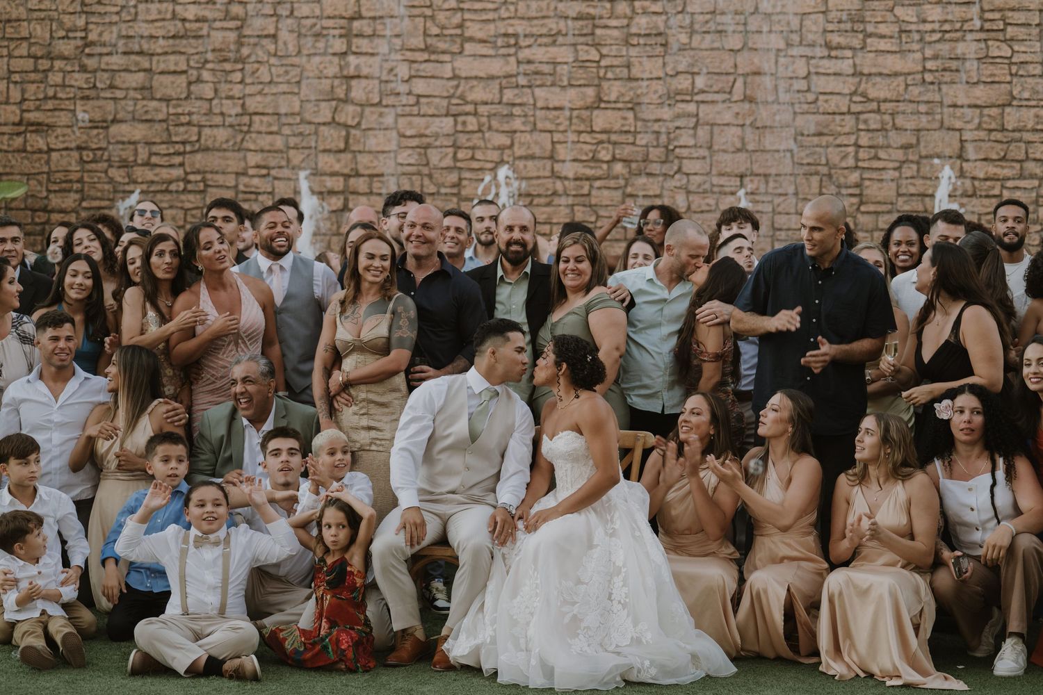 Bride and groom kiss surrounded by smiling wedding guests in a large group photo outdoors against a stone wall.