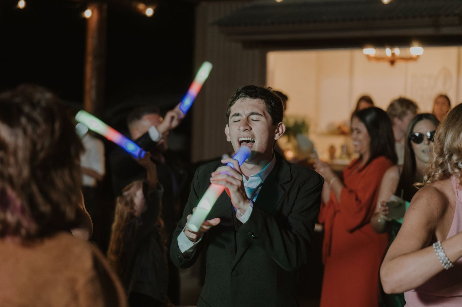 Singer performing with microphone at lively wedding reception while guests dance with glow sticks.
