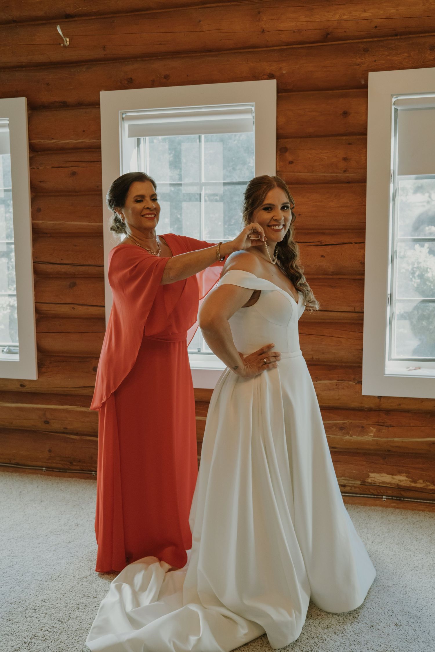 A bride in a white off-shoulder gown gets her necklace fastened by a woman in coral dress inside a rustic log cabin.