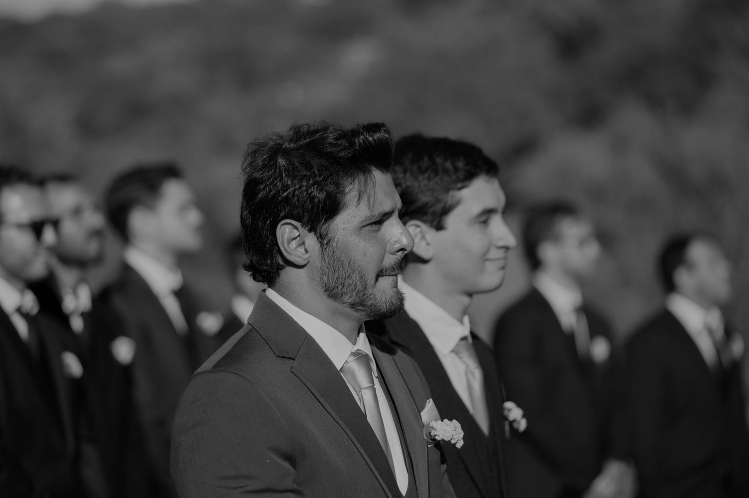 Black and white photo of groom smiling with groomsmen lined up at outdoor ceremony.