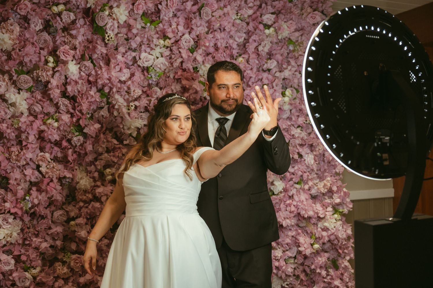 Bride and groom pose together in front of pink floral wall at wedding photo booth.
