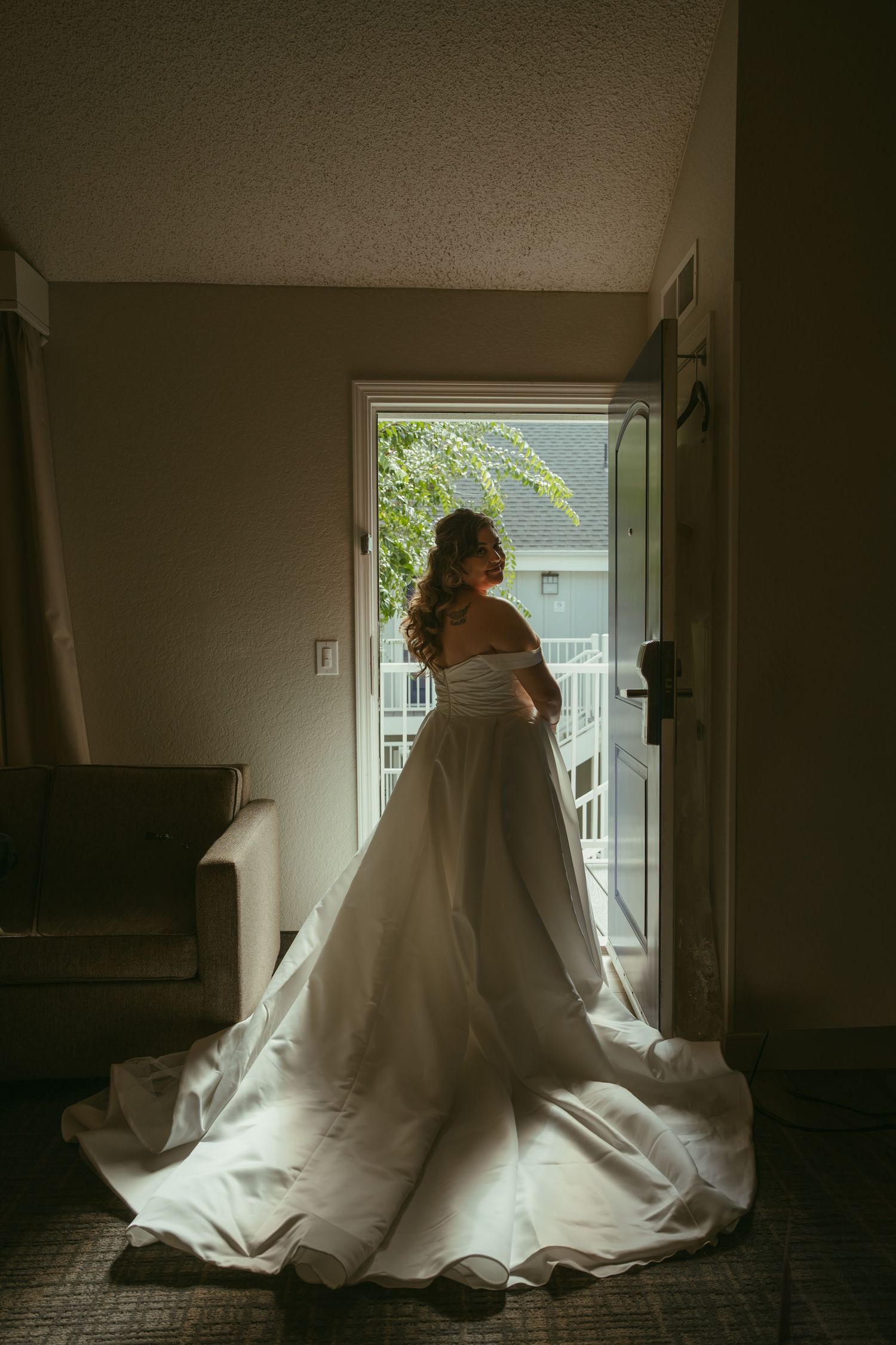 Bride in elegant off-shoulder white gown with long train standing in doorway, backlit by natural light.