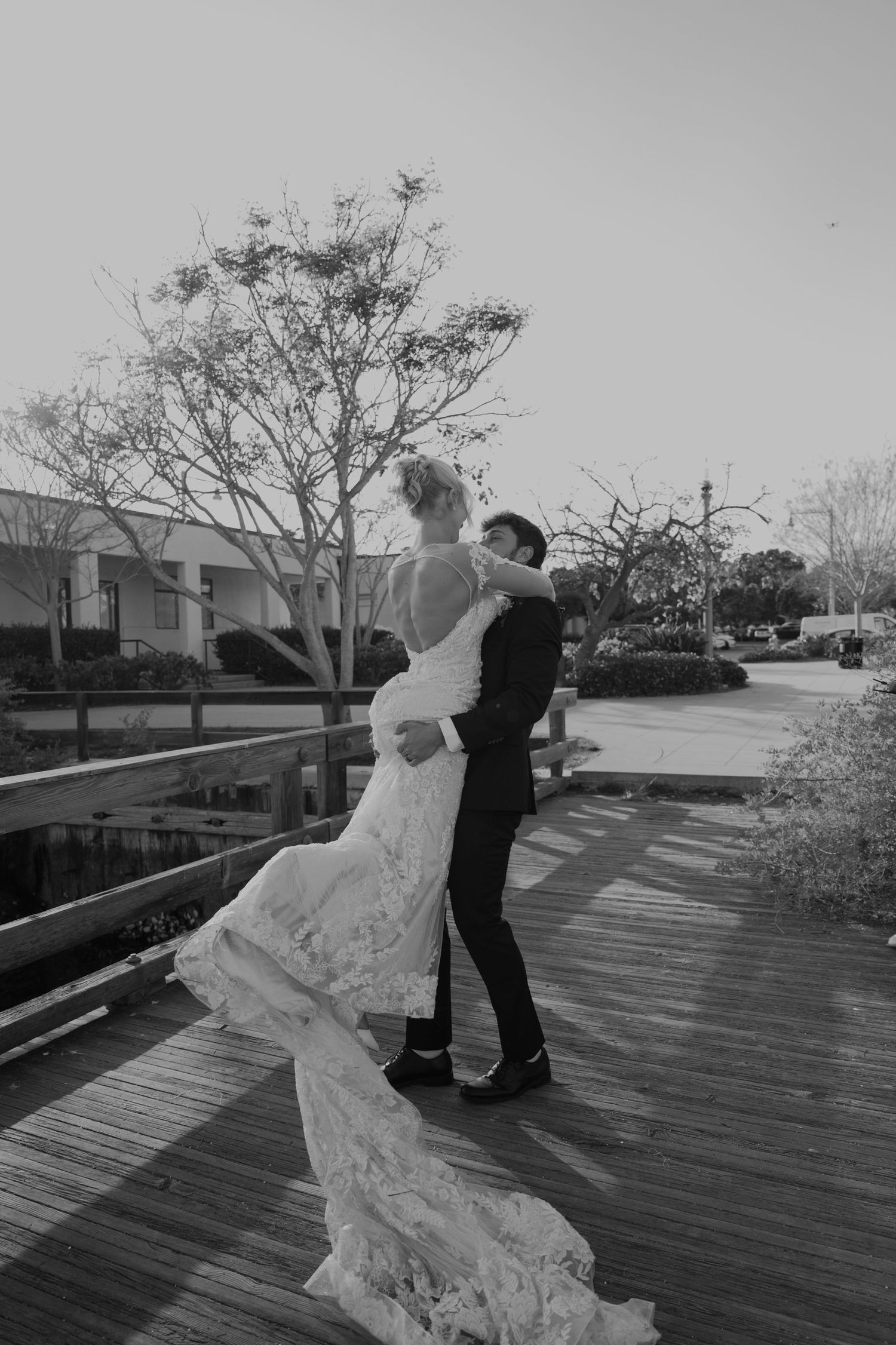Black and white photo of groom lifting bride in lace wedding dress on wooden deck outdoors.