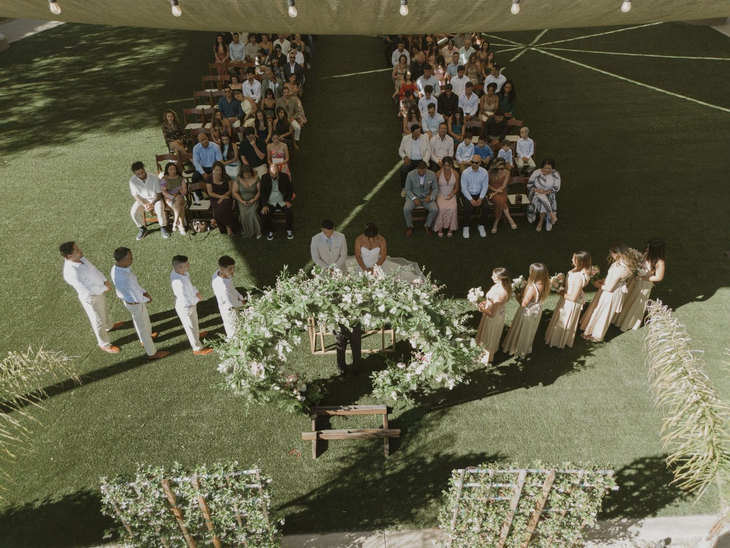 Aerial view of outdoor wedding ceremony with floral arch, wedding party, and guests seated on green lawn.