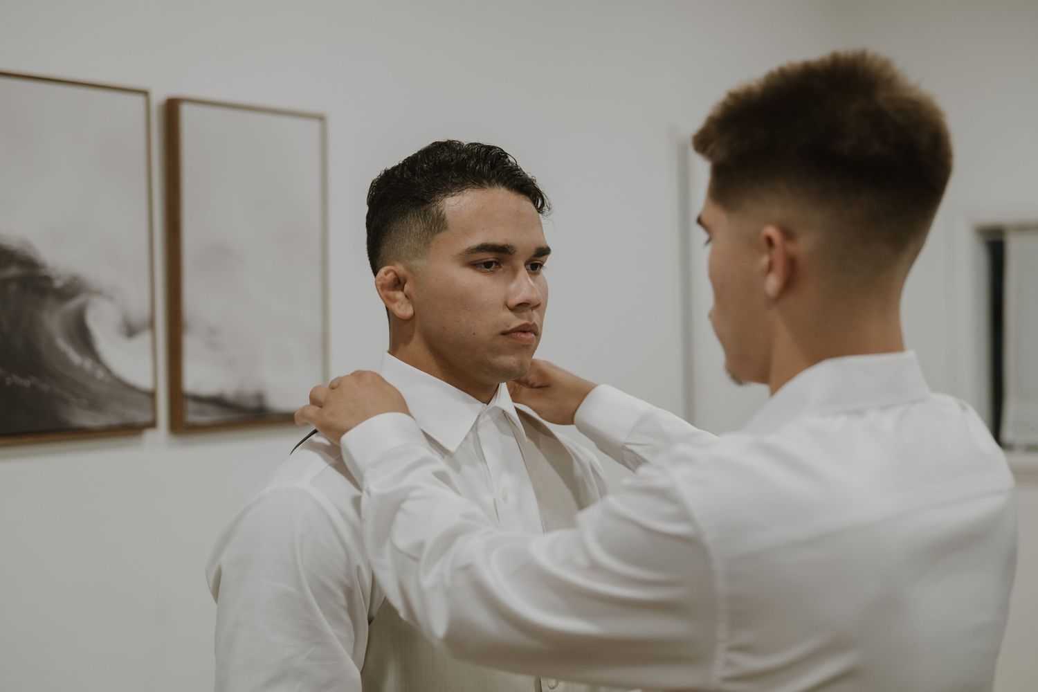 Groomsman helps adjust another mans white dress shirt collar before a wedding ceremony.