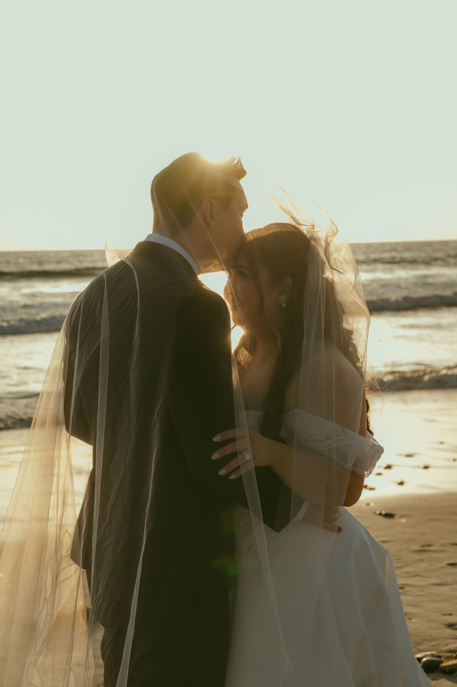 Bride and groom share a romantic moment on a sunlit beach, with her veil flowing in the breeze.