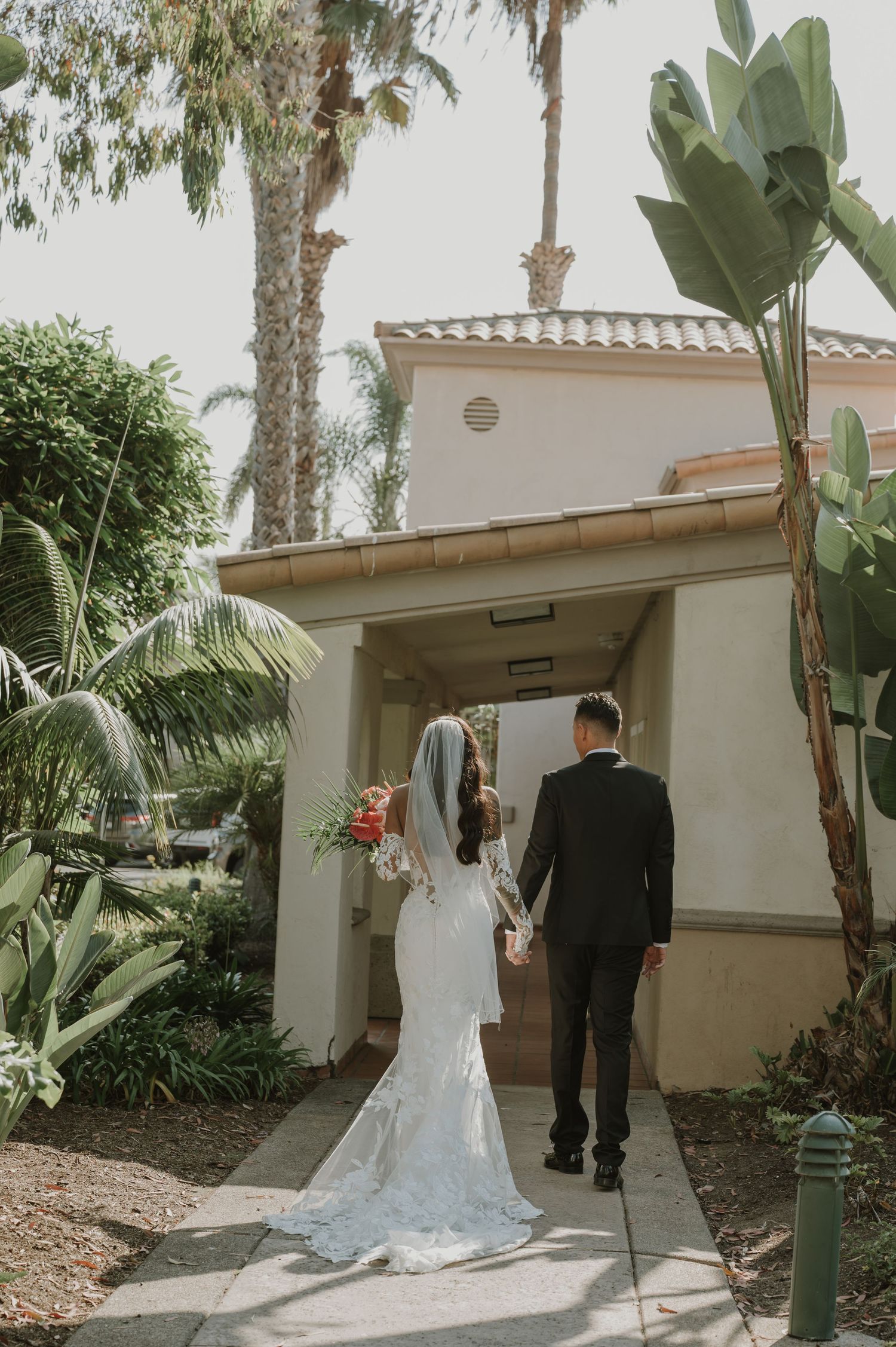 Wedding couple walking near Spanish-style buildings surrounded by tropical landscaping.