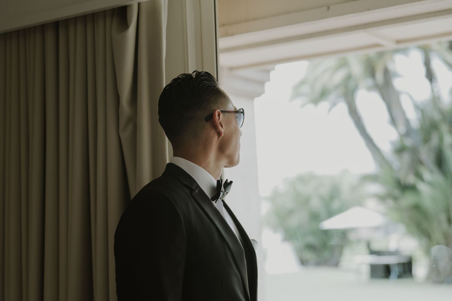 Side profile of groom in tuxedo and glasses looking out window.