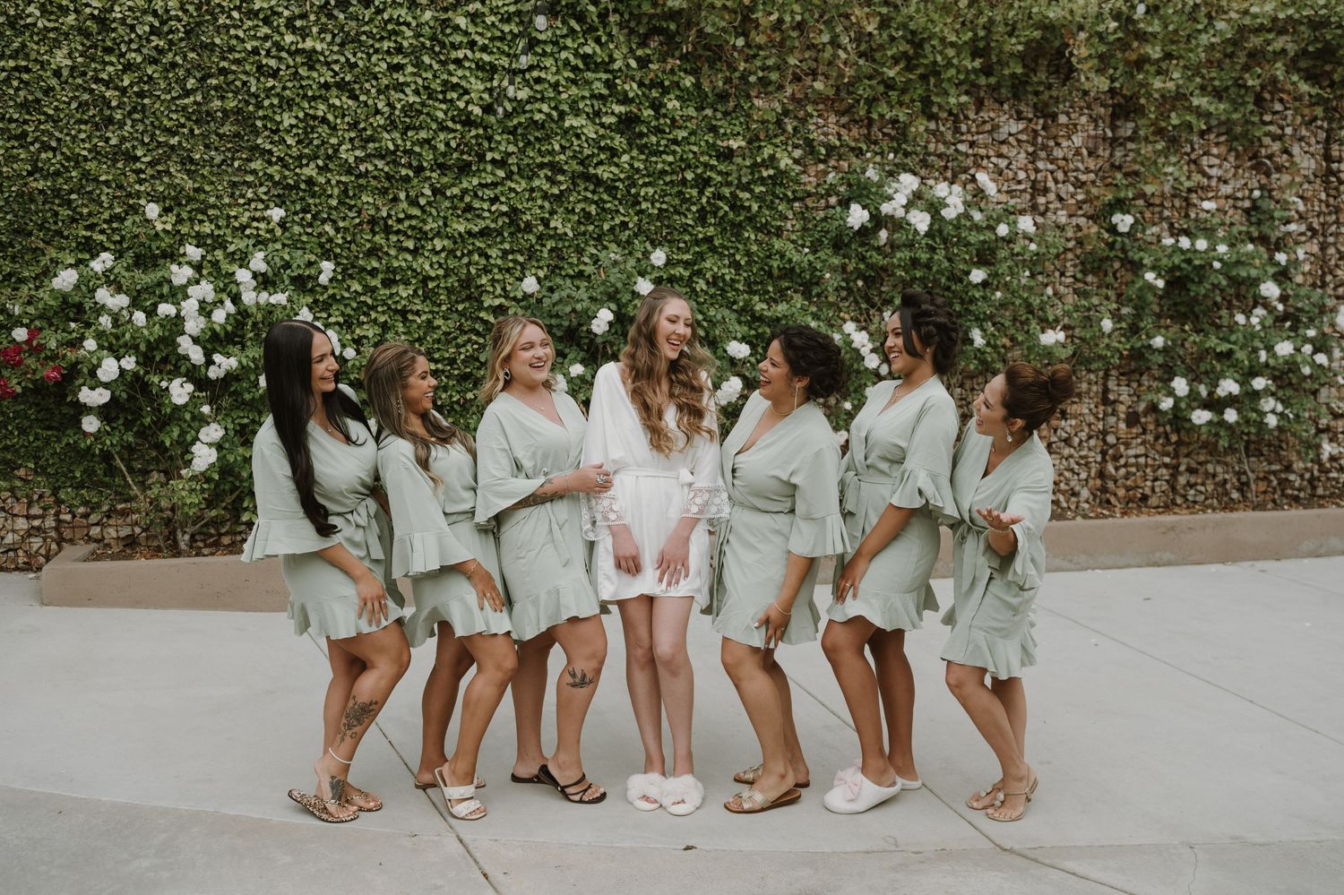 Bride in white robe laughing with six bridesmaids in sage green robes against an ivy wall with white flowers.