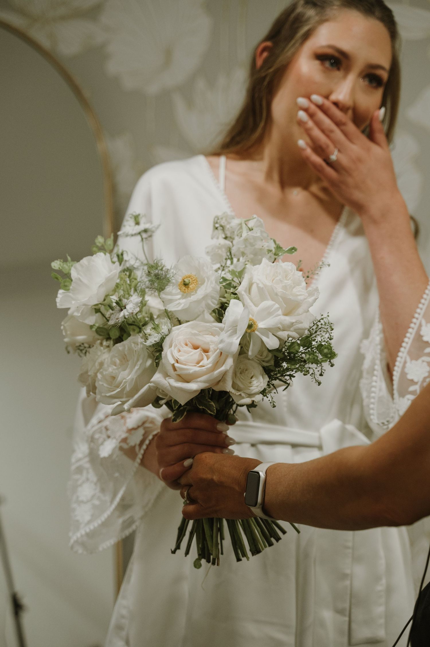 Emotional bride in white lace robe receiving her white rose and ranunculus bridal bouquet.