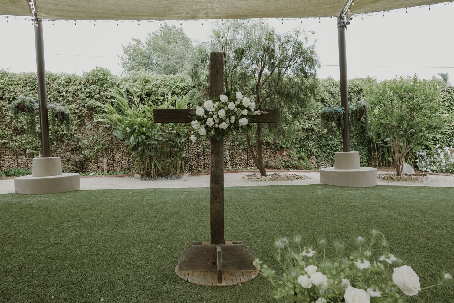 Rustic wooden cross adorned with white floral arrangement on green lawn at an elegant outdoor wedding ceremony.