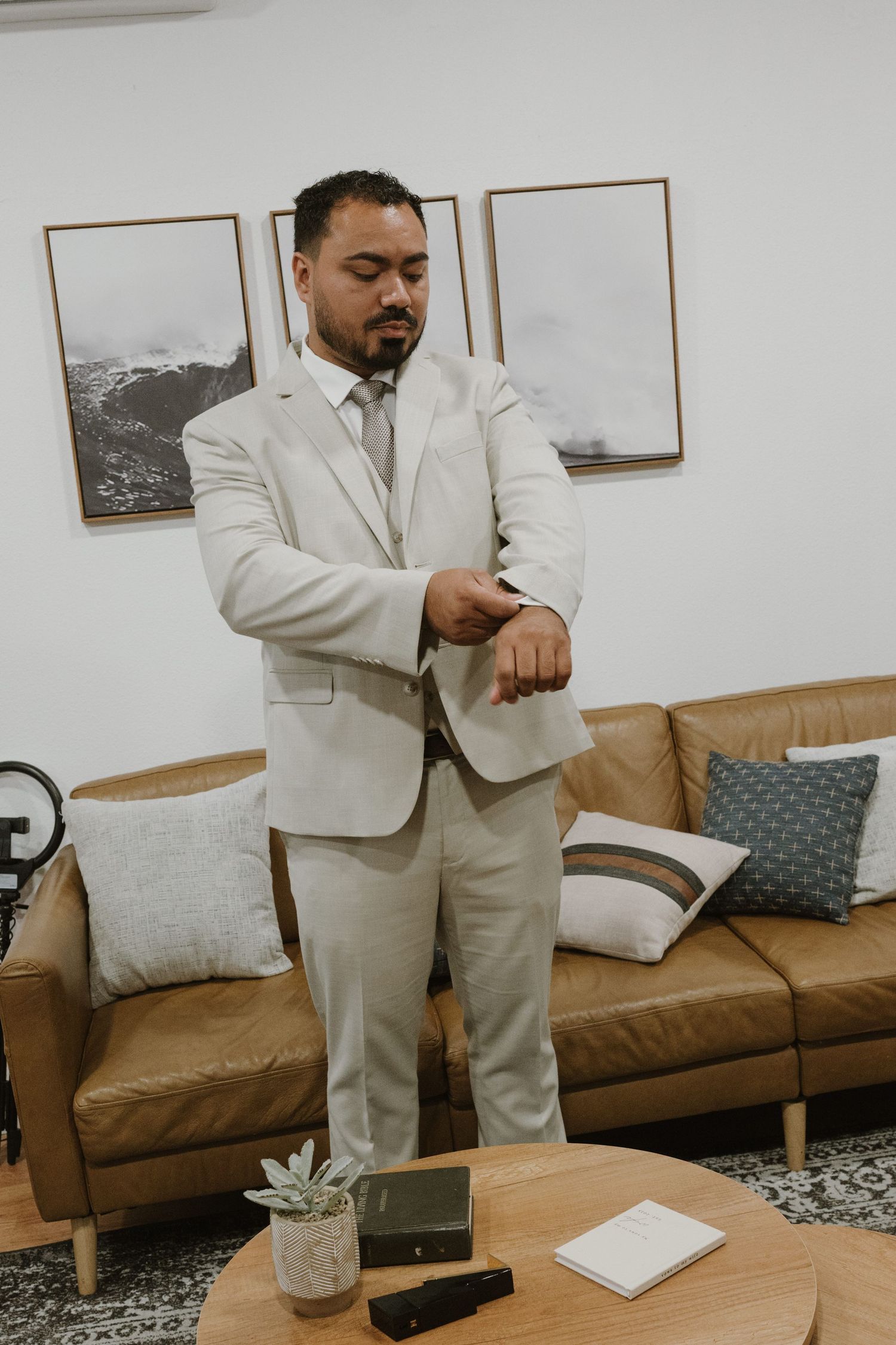 Man in light beige suit adjusting watch in modern living room with leather sofa and framed wall art.