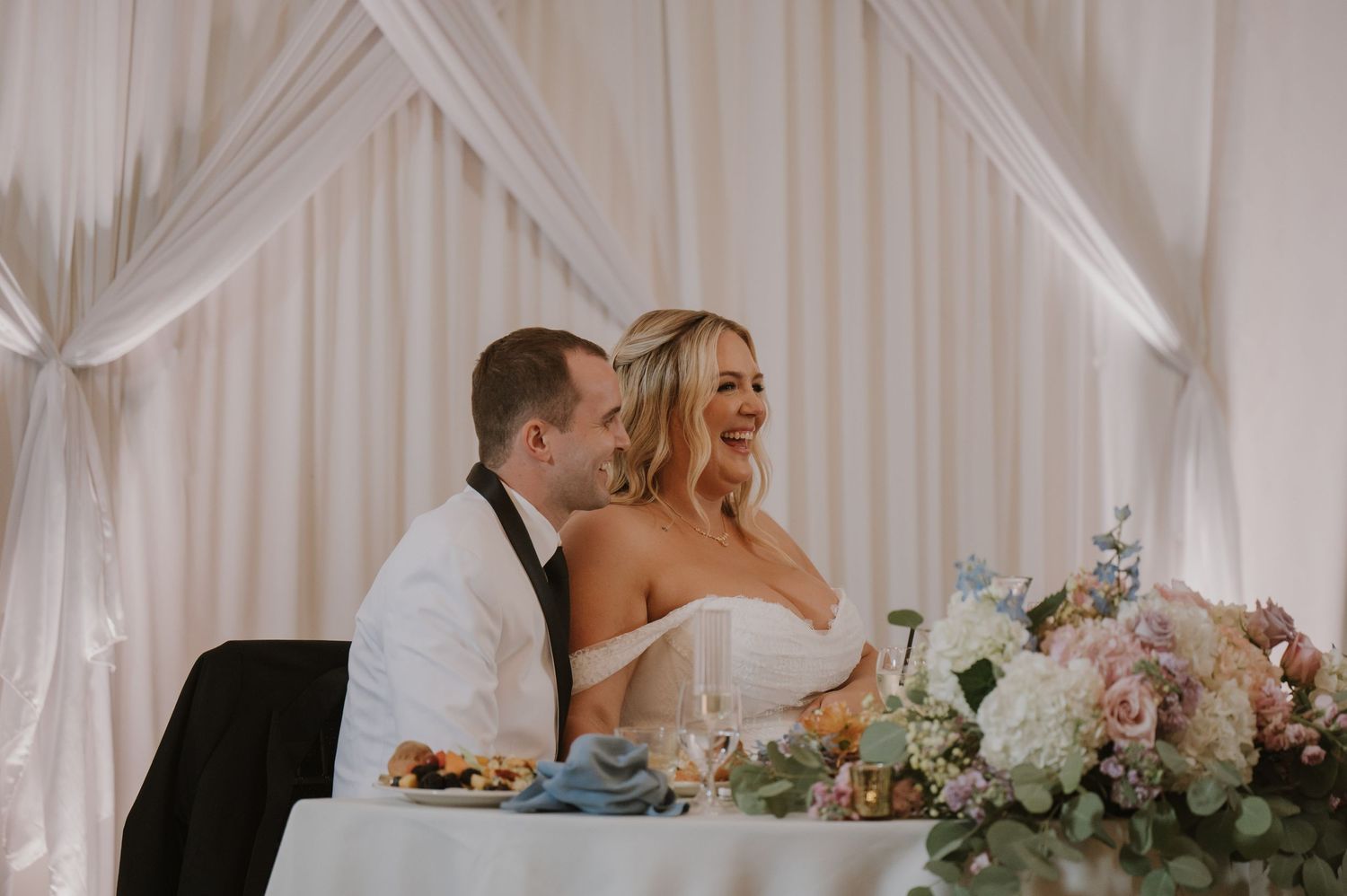 Bride and groom smiling at sweetheart table with floral centerpiece at elegant wedding reception.
