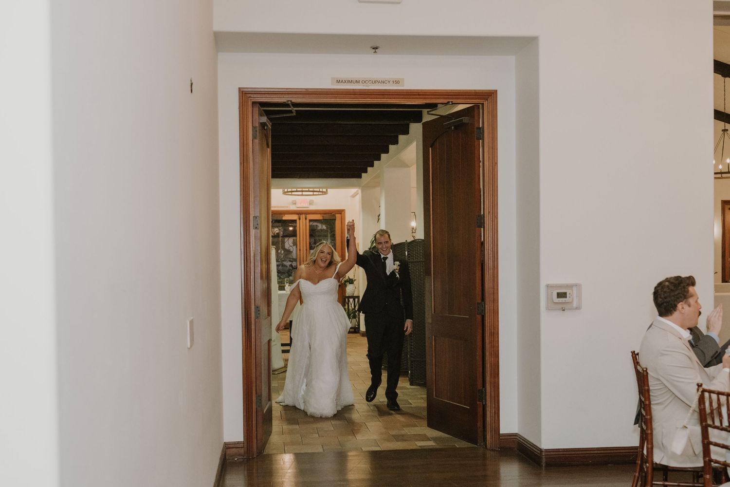 A bride and groom make their grand entrance through elegant wooden doors at their wedding reception venue.
