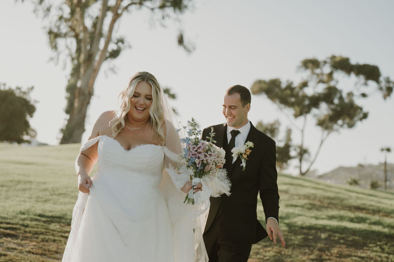 Smiling bride holding bouquet walks with groom on green hillside in sunny outdoor wedding portrait.