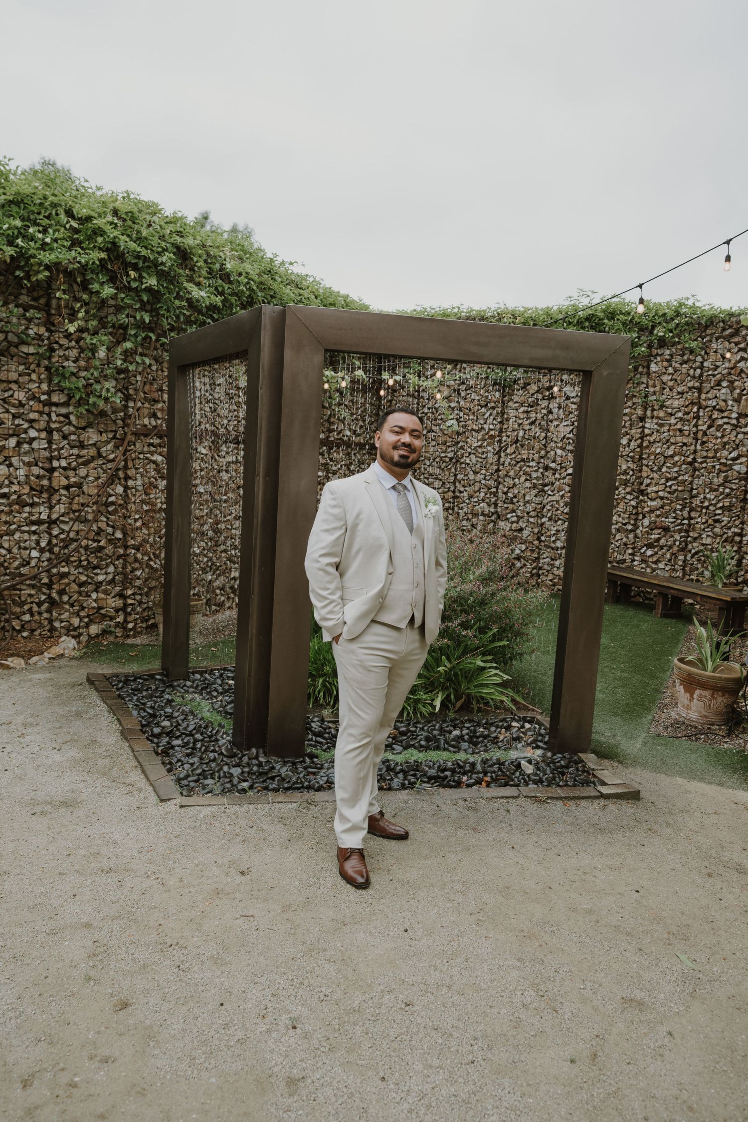 Groom in beige suit poses confidently before rustic wooden arch at outdoor wedding venue.