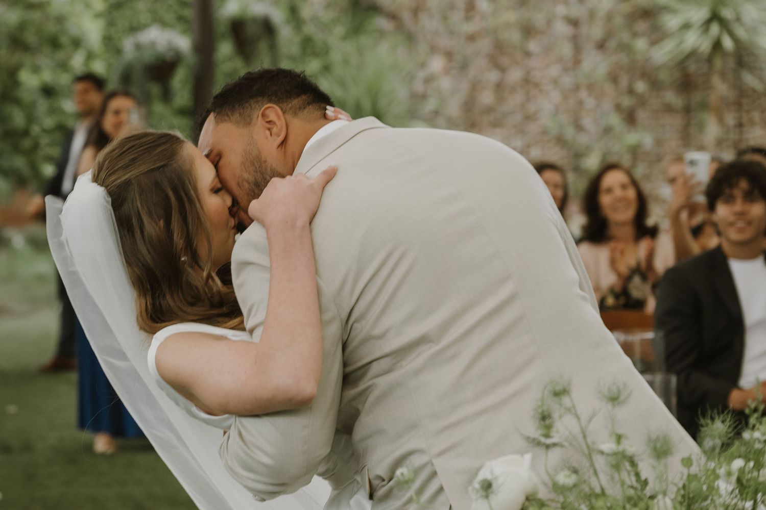A newlywed couple shares their first kiss during an outdoor garden wedding ceremony with guests applauding.