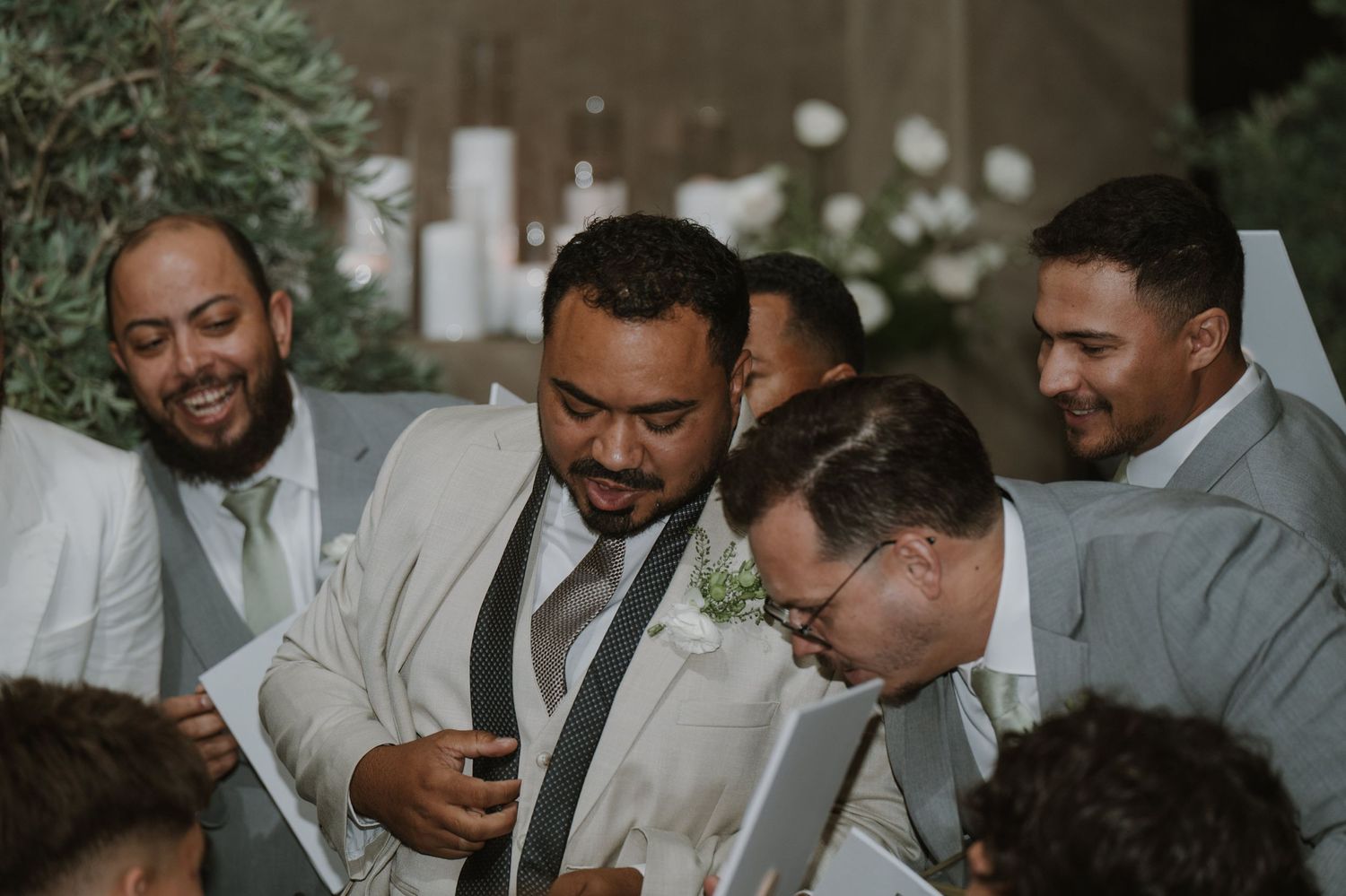 Groom in white suit laughing with groomsmen in gray suits during a joyful wedding ceremony moment.