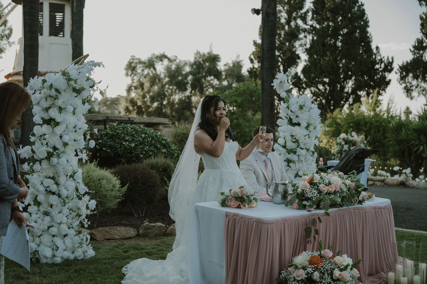 Bride and groom at outdoor wedding ceremony beneath white floral arch at dusk.