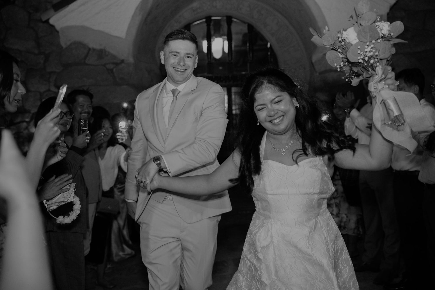 Smiling bride and groom making a grand exit through cheering wedding guests in black and white.