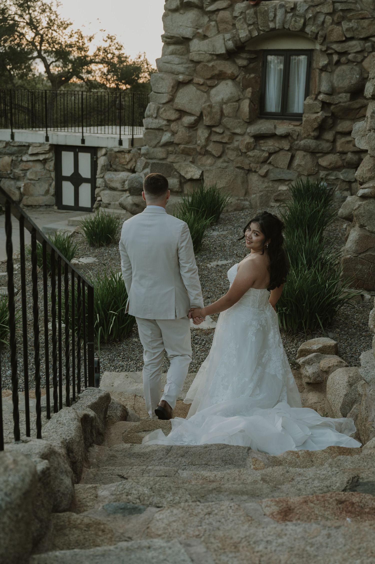 Bride in white gown and groom in light suit hold hands walking up stone steps.