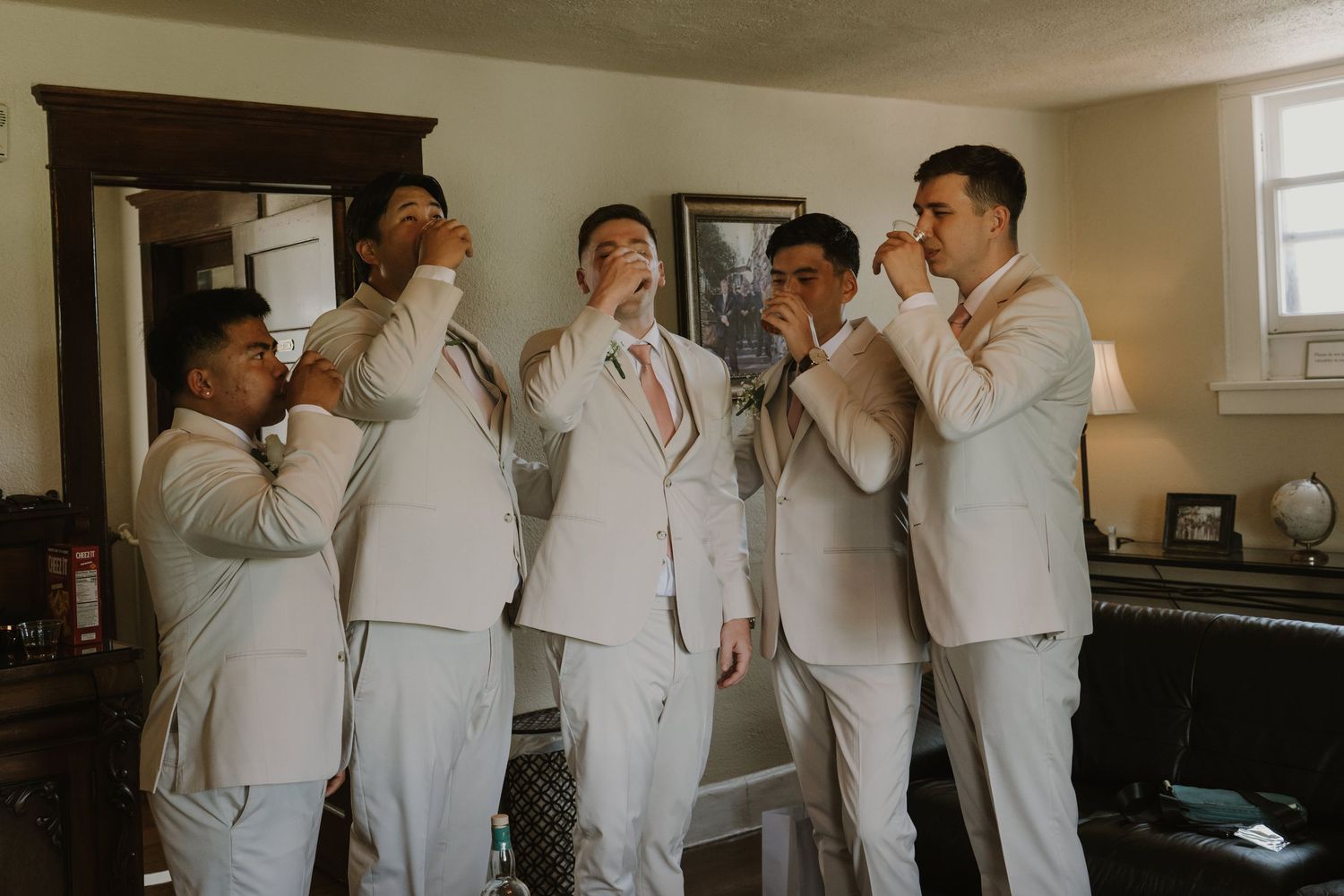 Five groomsmen in matching cream suits adjusting their ties together while getting ready for a wedding ceremony.