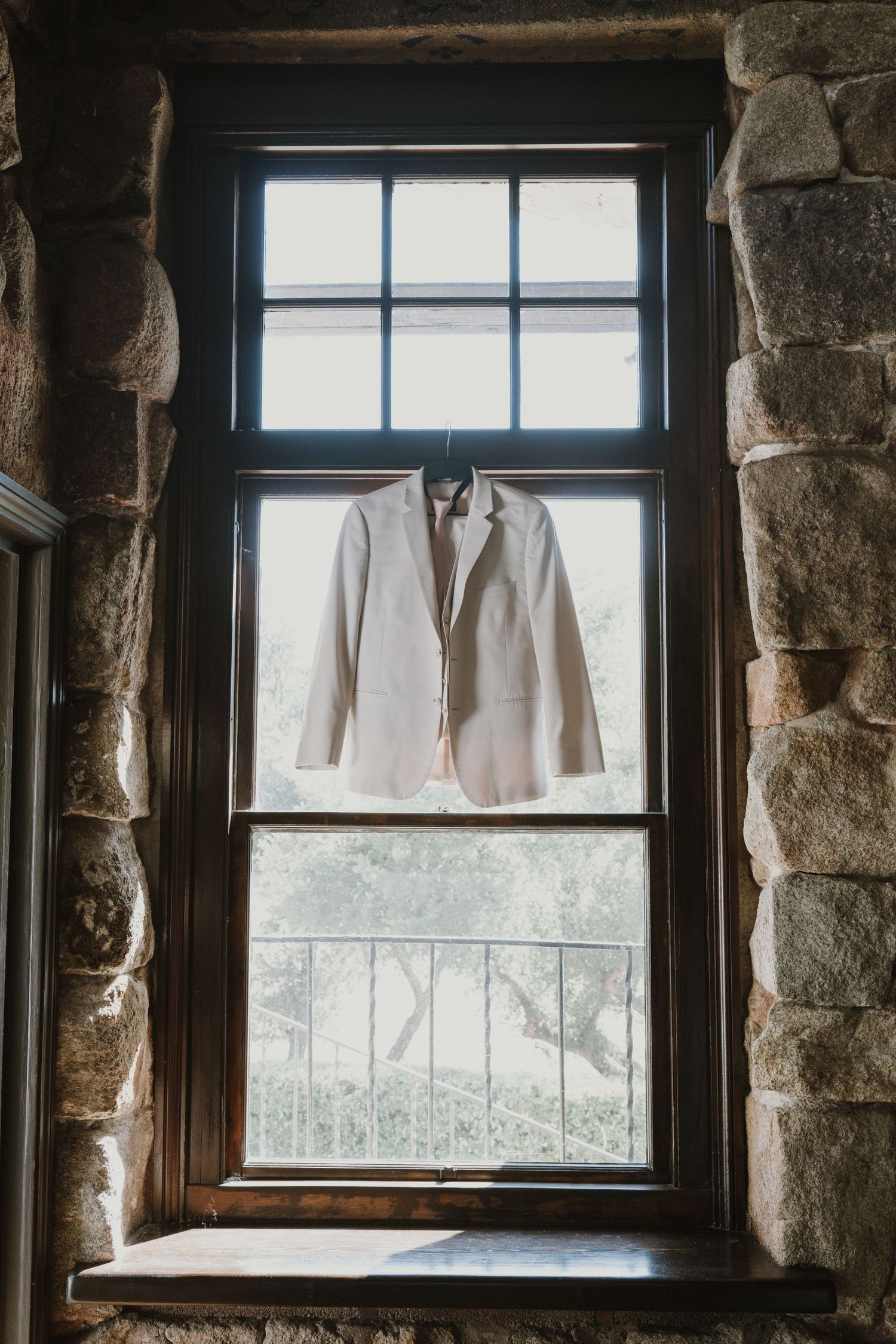 A white wedding suit jacket hanging on a hanger in a rustic stone window with natural light.
