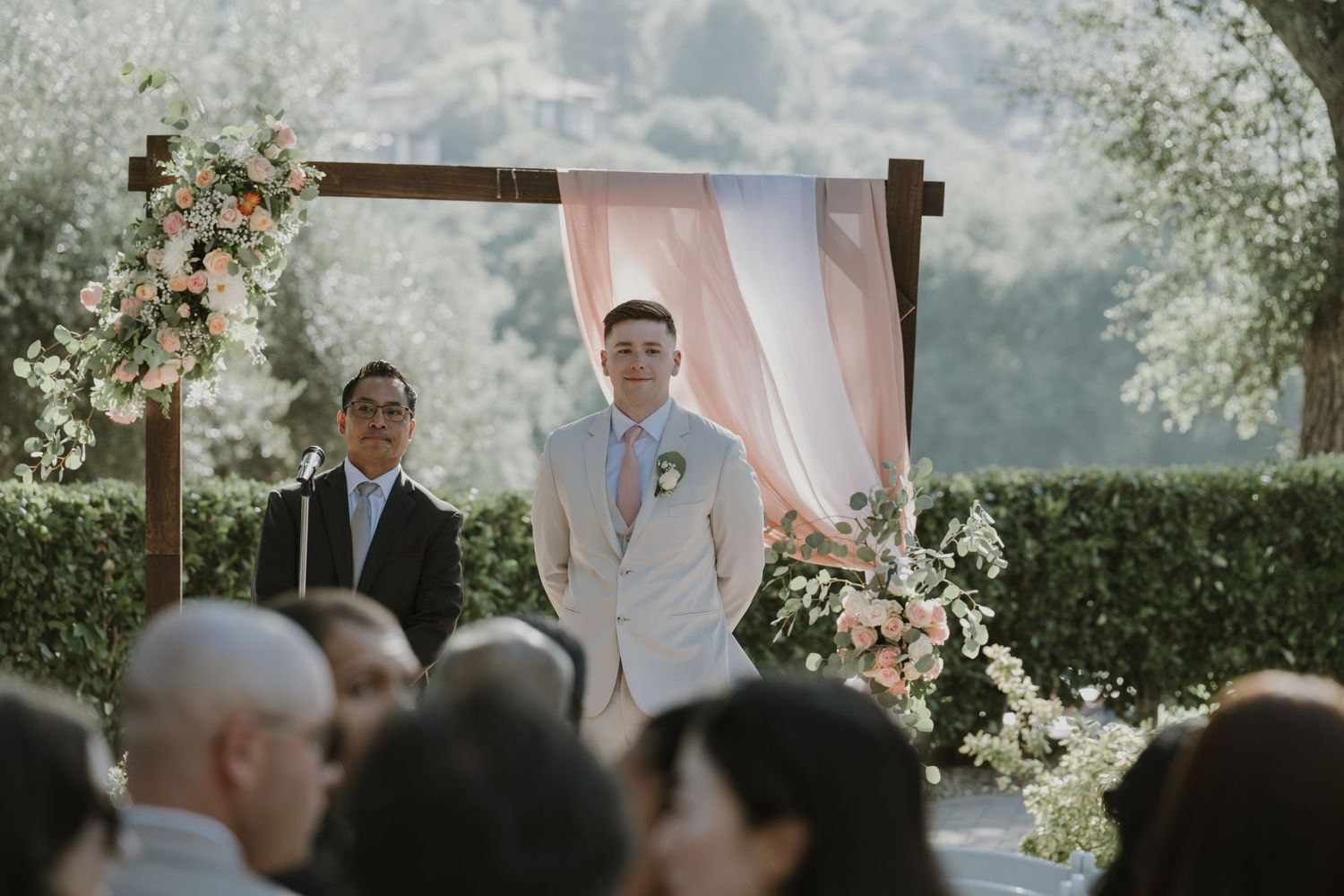 Groom in beige suit stands at floral wedding altar with pink draping and lush greenery backdrop.