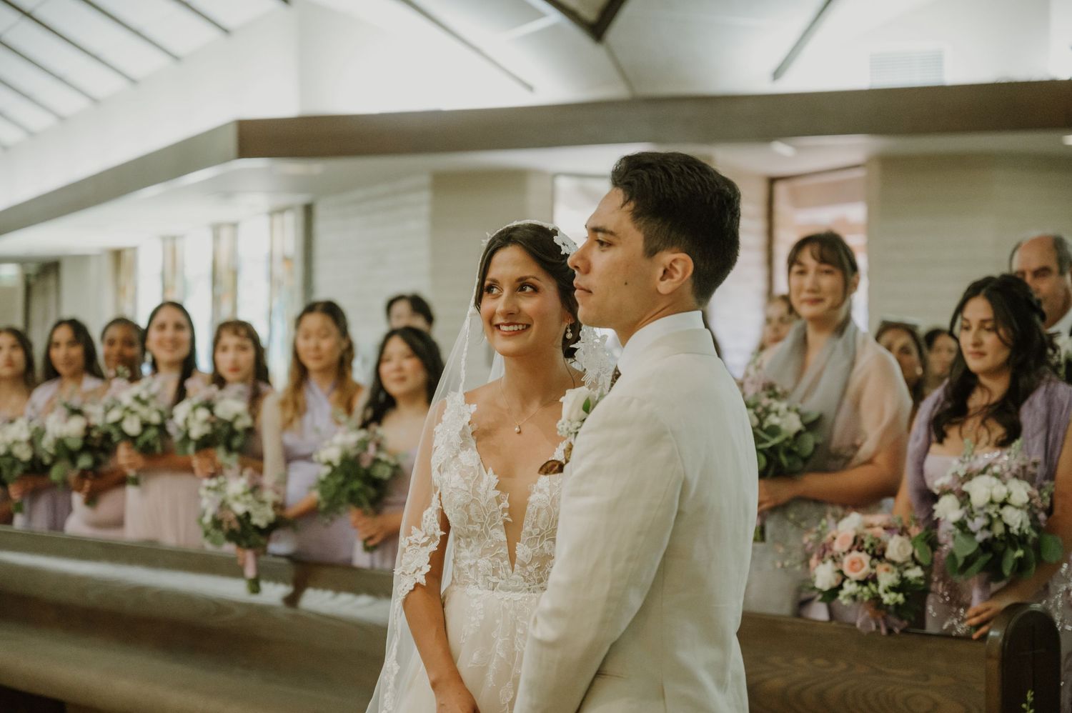 Bride and groom stand at church altar smiling, bridesmaids in pink dresses holding floral bouquets behind them.