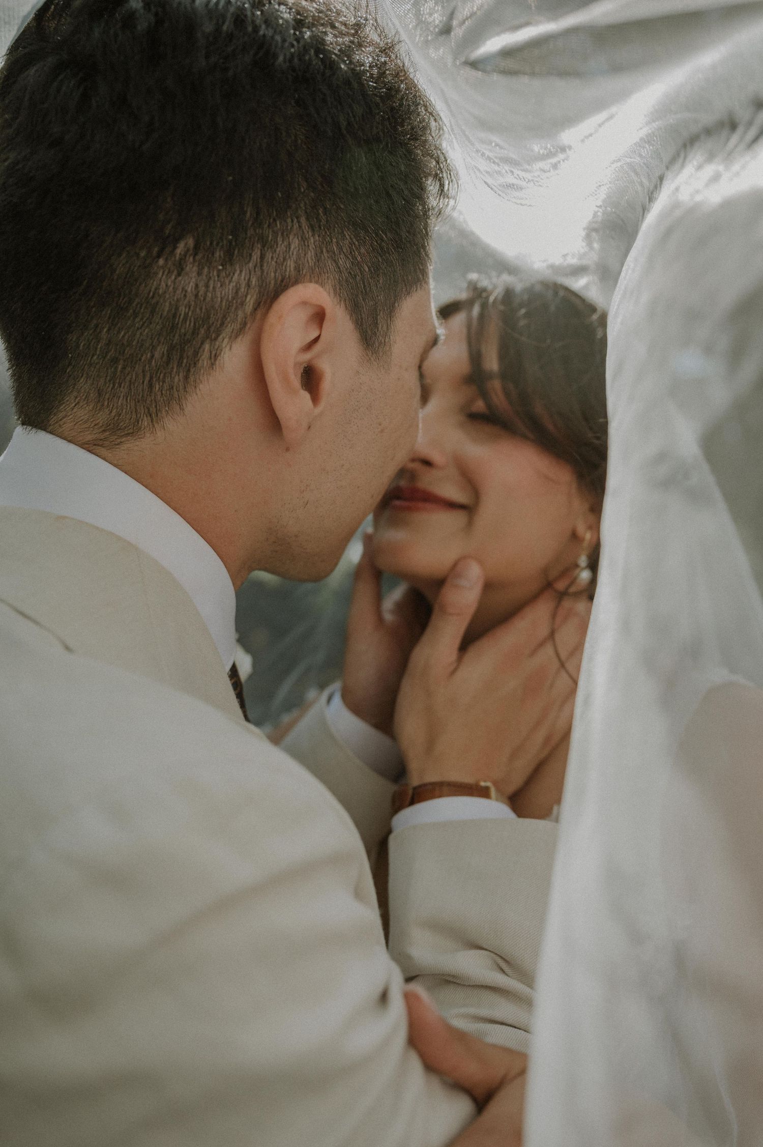 Bride and groom share an intimate moment under a white veil, nearly kissing.