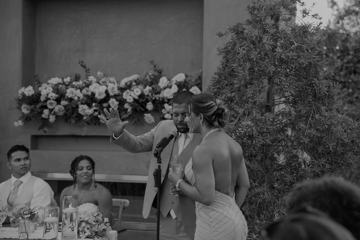 Bride and groom share a toast while giving speech at outdoor wedding reception in black and white.