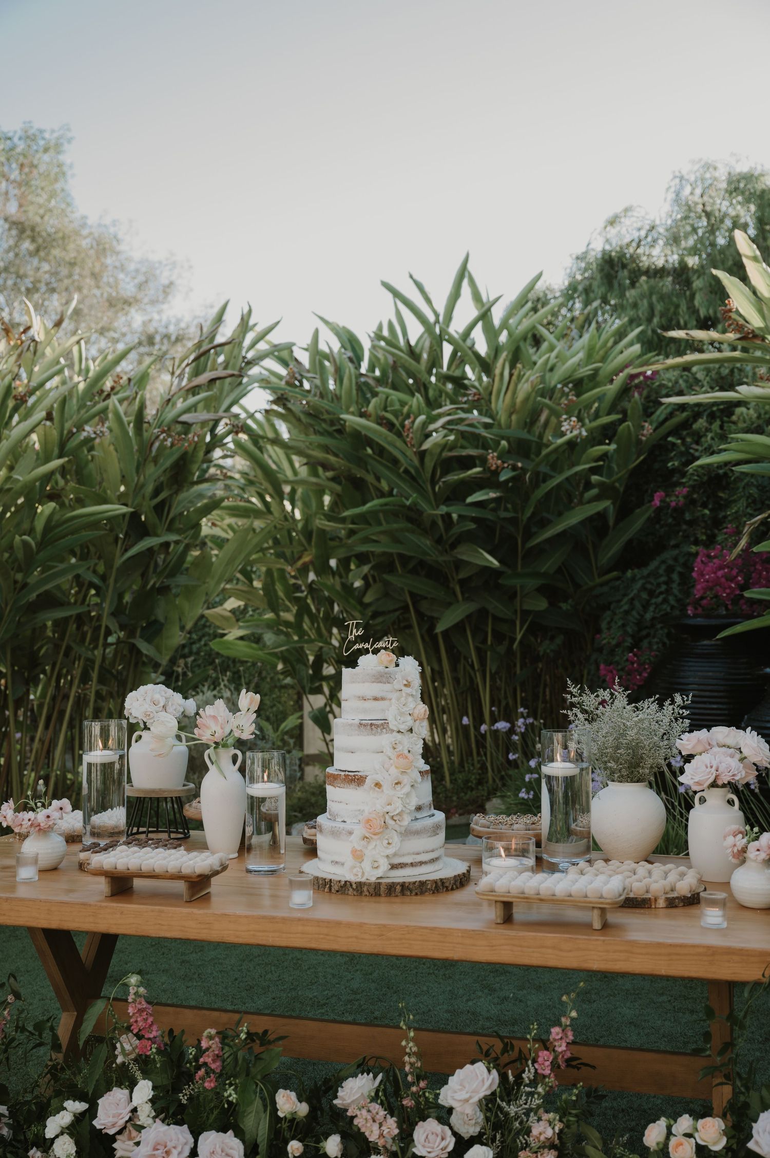 Elegant outdoor wedding dessert table with tiered naked cake, white florals, and tropical greenery backdrop.