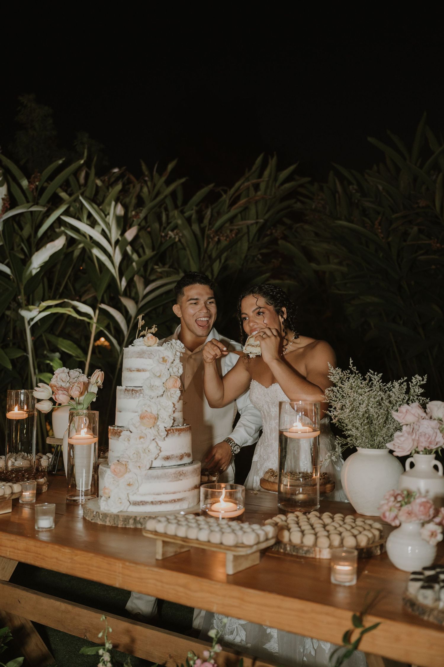 Newlyweds joyfully cutting their white floral wedding cake at a candlelit dessert table.