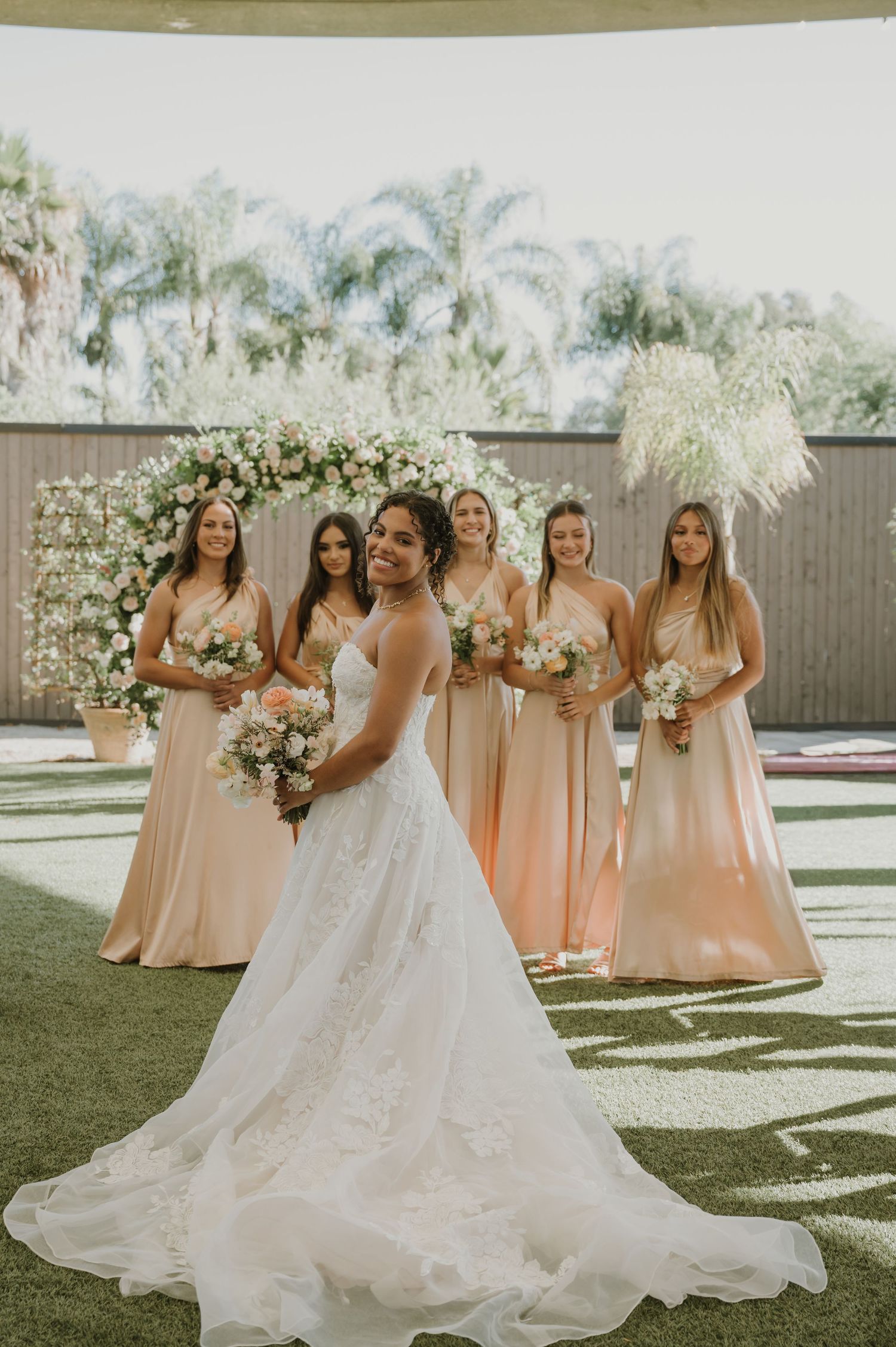 Bride in white lace gown poses with four bridesmaids in blush dresses holding bouquets before floral arch.