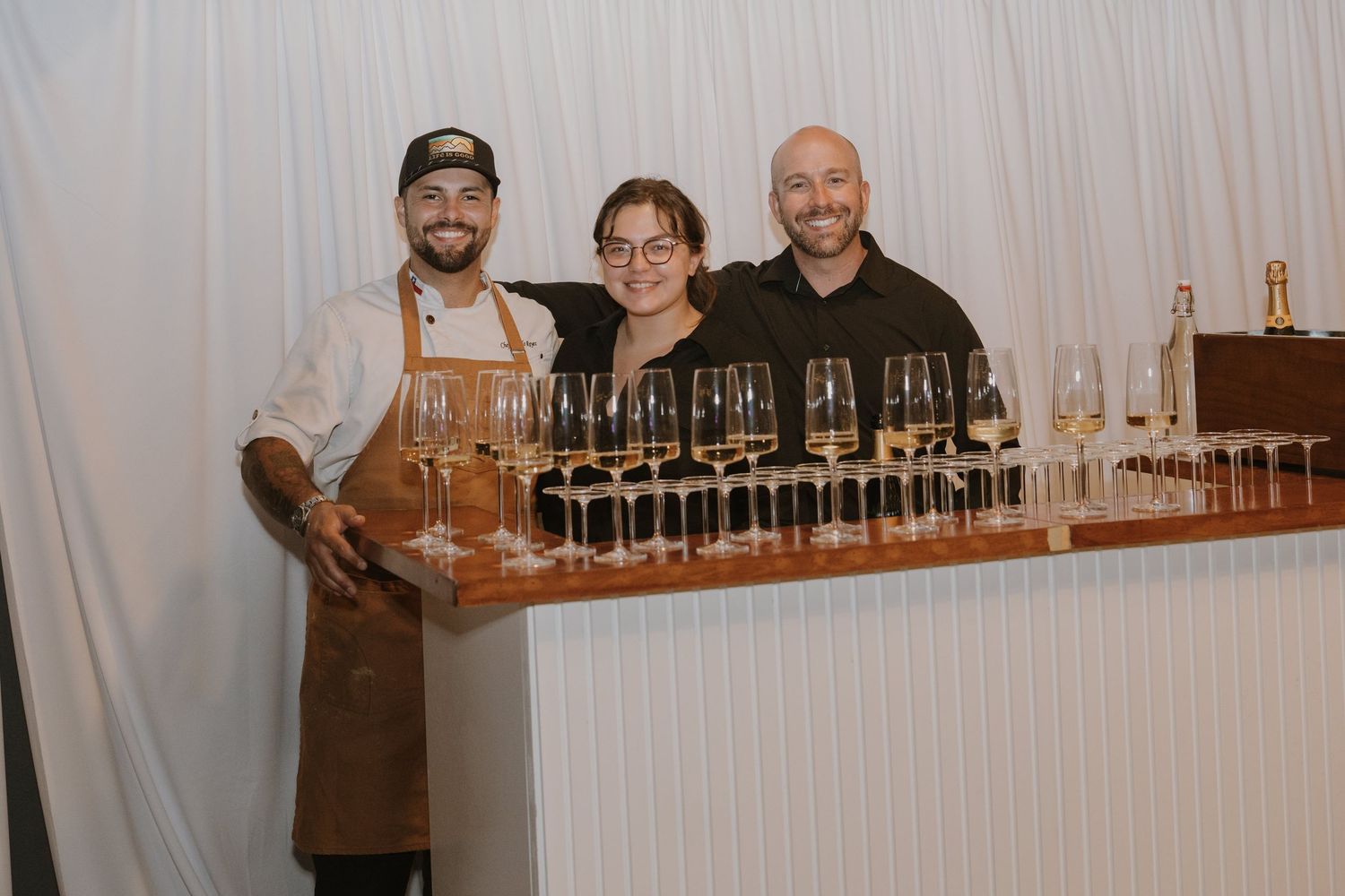 Three smiling staff members pose behind a bar with wine glasses at an elegant catered event.