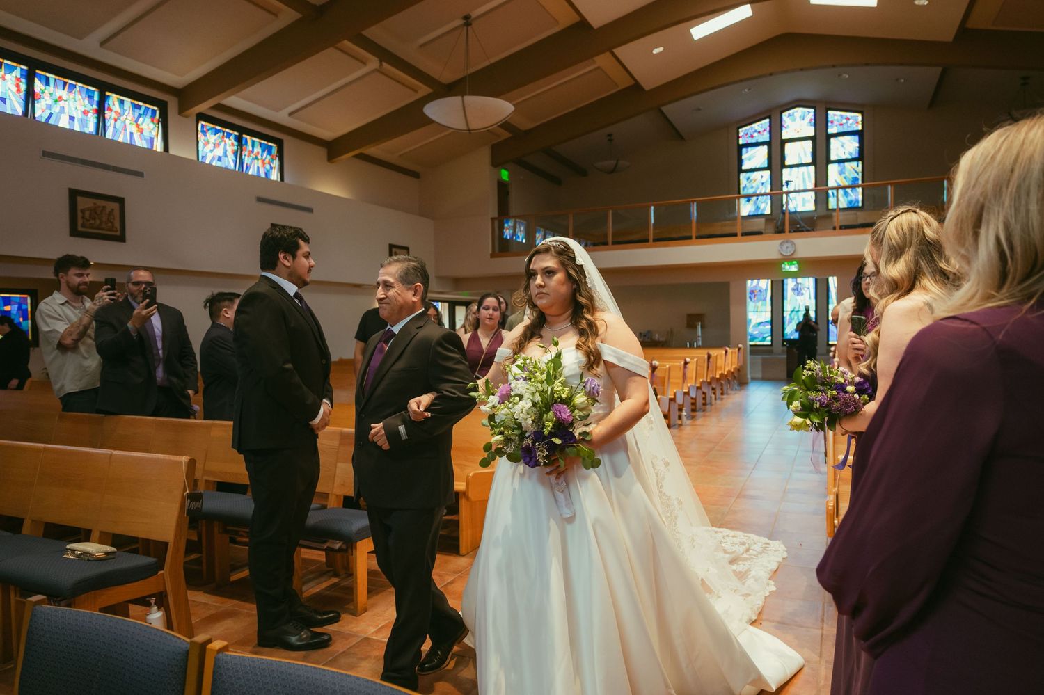 Bride in white gown walks down church aisle with escort, holding floral bouquet, guests standing in wooden pews.