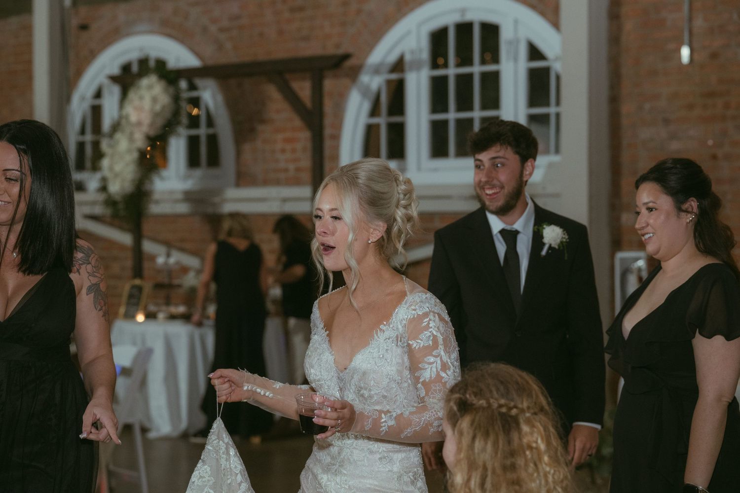Bride in lace gown laughing with guests on wedding reception dance floor.