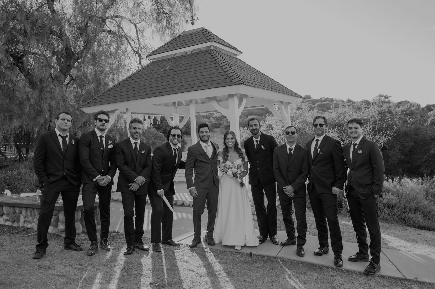 Black and white photo of bride, groom, and groomsmen posing near gazebo in outdoor wedding venue.
