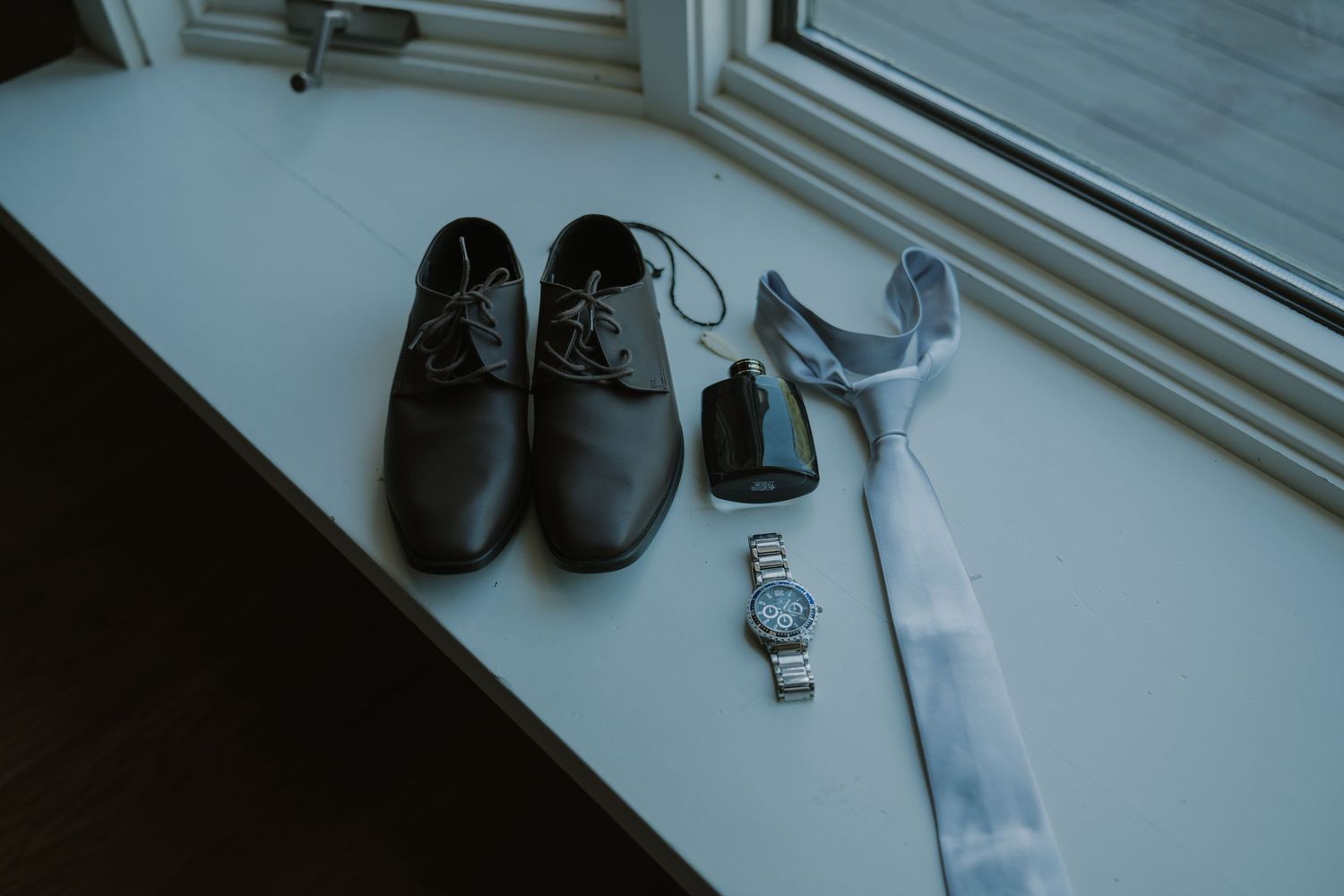 Brown leather dress shoes, watch, cologne, and light blue tie arranged on a windowsill for a groom wedding preparation.