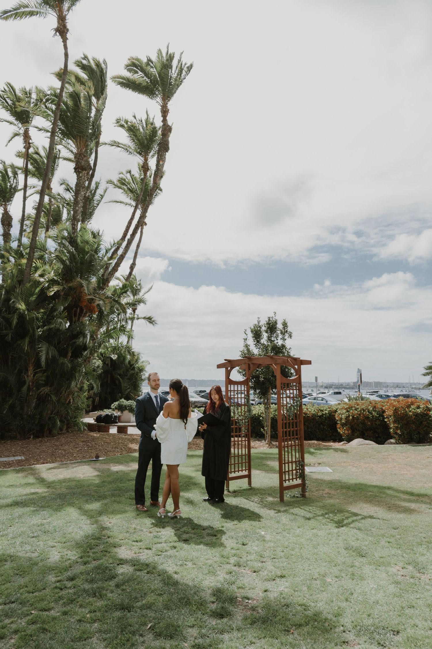 Outdoor wedding ceremony setup with palm trees and wooden arch against cloudy sky.
