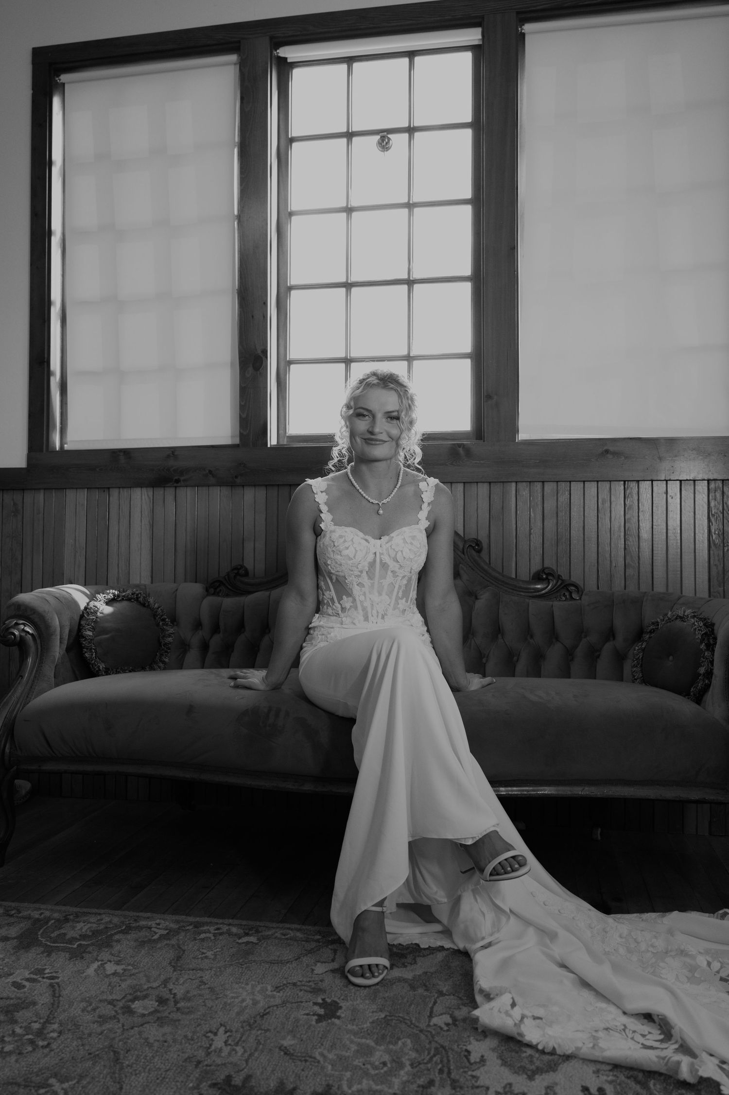 A bride in a white lace gown reclines elegantly on a tufted couch in a sunlit room.