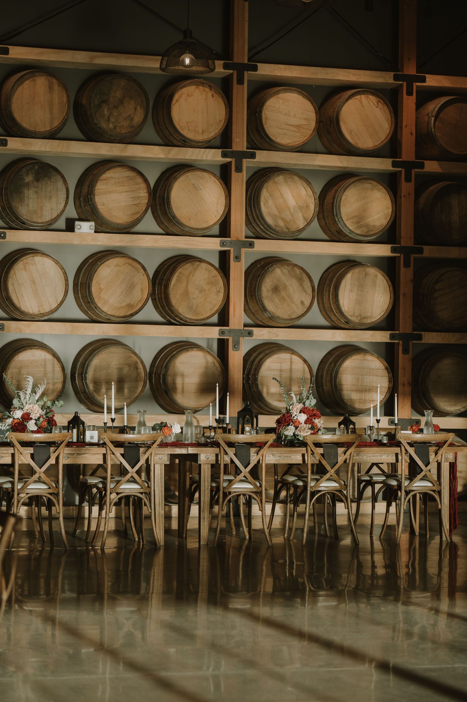 Long banquet tables set up in front of stacked wine barrels in an industrial-chic winery space.
